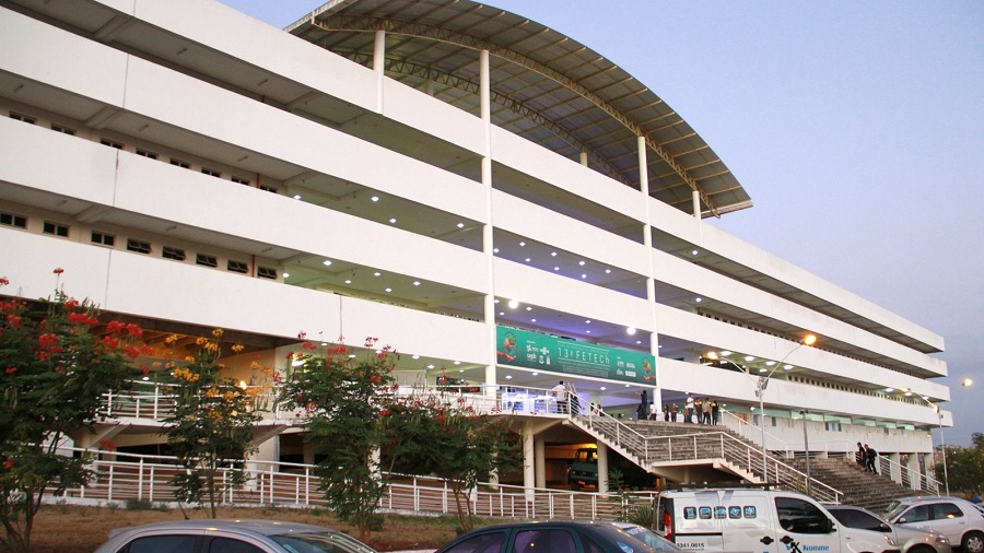 Fotografia em vista diagonal da fachada frontal do prédio do campus da UEPB, em Campina Grande, que é um edifício horizontal de quatro andares, branco, com uma telha metálica curva sobre a cobertura.