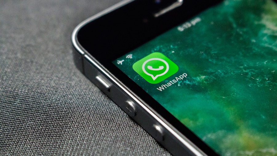 Fotografia em plano fechado e vista superior que mostra o canto de um celular, onde vê-se o ícone do Whatsapp sobre um fundo de tela de cor verde marmorizado; o aparelho está sobre um tecido de cor cinza.