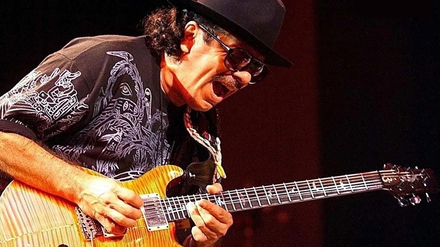 Fotografia em primeiro plano de Santana, com um chapéu preto, óculos escuros e a boca aberta, enquanto sola em uma guitarra, com corpo de cor bege e listras laranjas, de sua própria marca, e um fundo escuro.