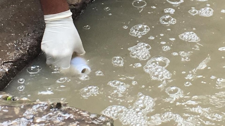 Fotografia que mostra uma mão calçada com luva branca que mergulha um frasco branco em uma água turva de esgoto, com várias bolhas em sua superfície, margeada por concreto, que pode se ver nas partes esquerda e inferior da imagem.
