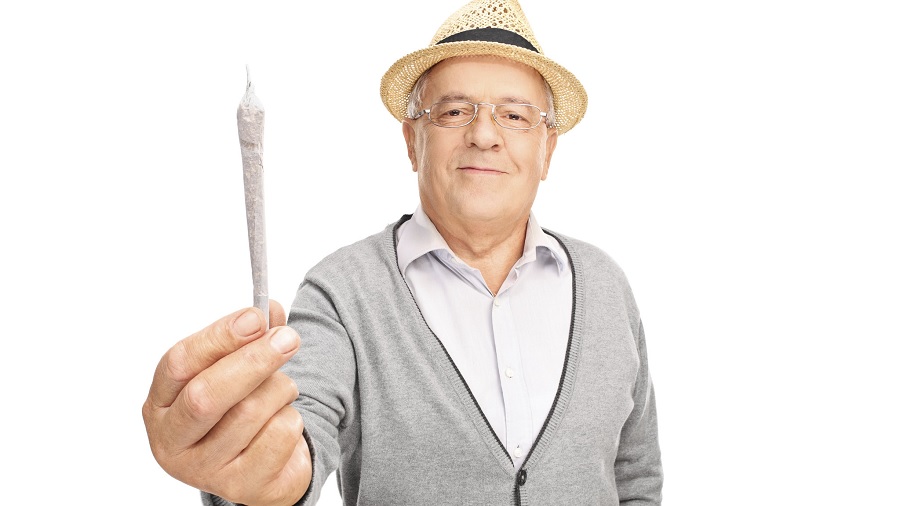 Fotografia em primeiro plano de um homem de meia-idade, usando um chapéu panamá de palha, óculos e um casaco de cor cinza-claro, sobre uma camisa branca, e segurando um cigarro de maconha, na vertical, diante da câmera, com um fundo branco.