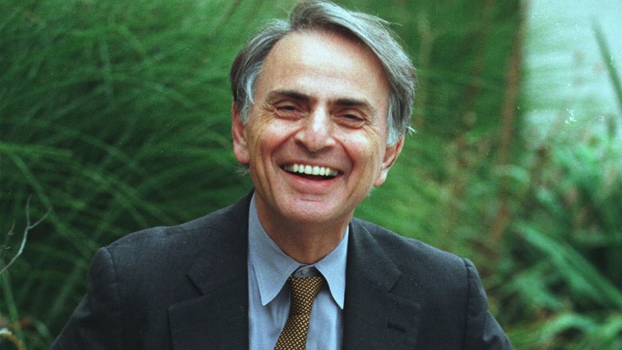 Fotografia em primeiro plano que mostra Carl Sagan com um sorriso aberto, usando paletó cinza-escuro e camisa azul-claro, e, ao fundo, desfocado, uma vegetação.