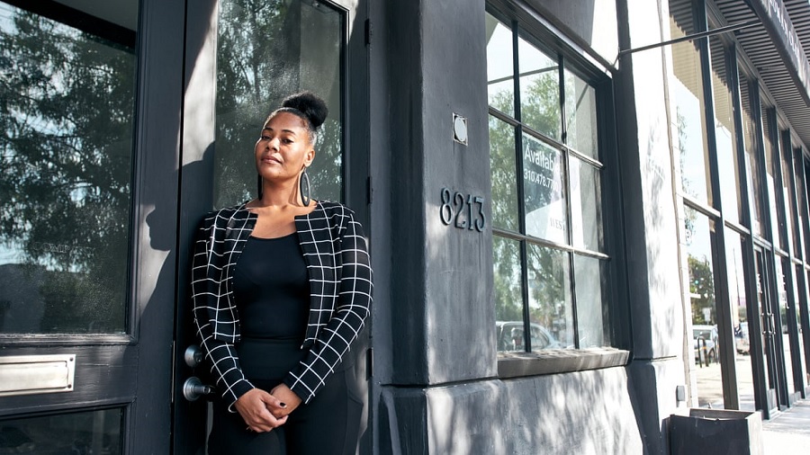 Fotografia em primeiro plano de Lanaisha Edwards, vestindo uma roupa de cor preta, com as mãos unidas à frente do corpo e encostada na porta do espaço para loja de cannabis que alugou, em Beverly Grove.