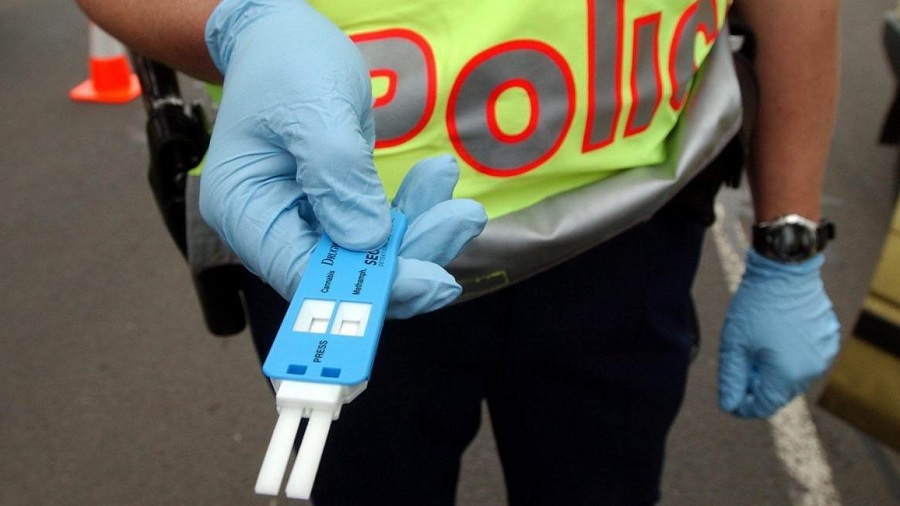 Fotografia em vista superior que mostra parte do corpo de um policial que está usando luvas azuis e segurando um drogômetro, de mesma cor, com duas fitas brancas na ponta.