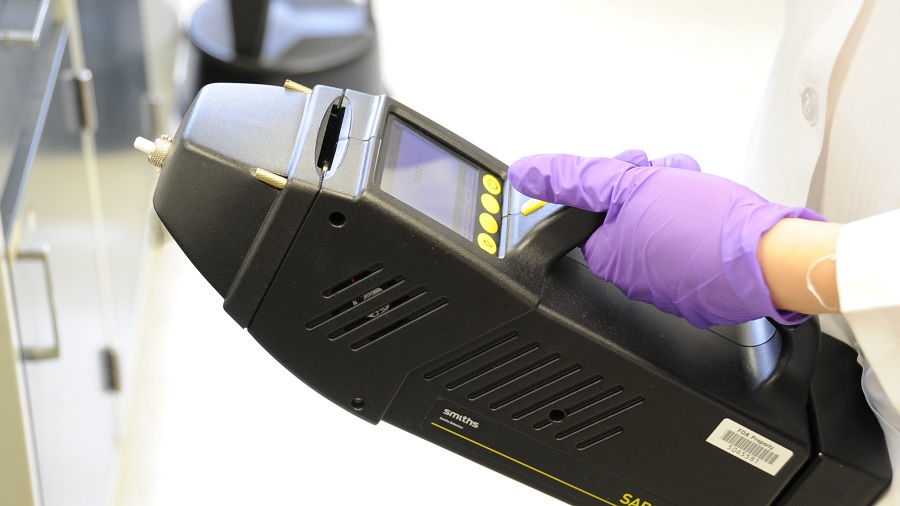 Foto que mostra um aparelho portátil de realização de diagnóstico através de espectroscopia Raman, em formato retangular e cor preta, e mão da pessoa que o segura usando luva lilás. Imagem: FDA | Flickr.