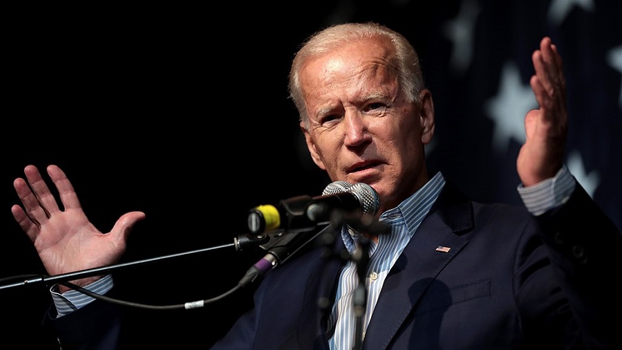 Fotografia em primeiro plano de Joe Biden, com os braços flexionados para cima, as mãos abertas e os olhos cerrados, enquanto fala ao microfone, e um fundo escuro. Foto: Gage Skidmore | Creative Commons.