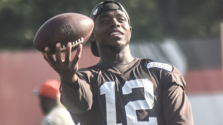 Fotografia que mostra, em primeiro plano, o jogador Josh Gordon vestido com um uniforme do Browns, de cor preta e com o número “12” em branco, e segurando uma bola de futebol americano à sua frente e um fundo desfocado.