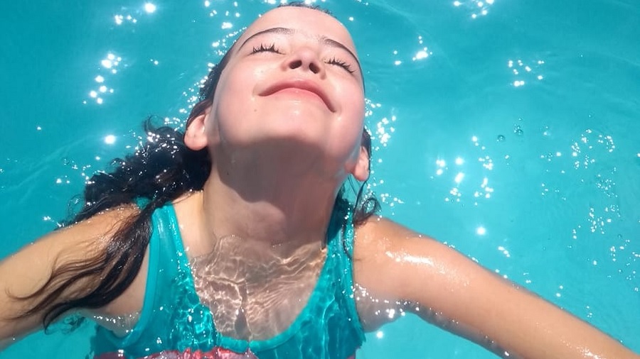 Fotografia em vista superior que mostra Caroline, do peito pra cima, dentro de uma piscina, com a cabeça para fora, a face voltada para cima, os olhos fechados e um sorriso nos lábios. Foto: arquivo pessoal.