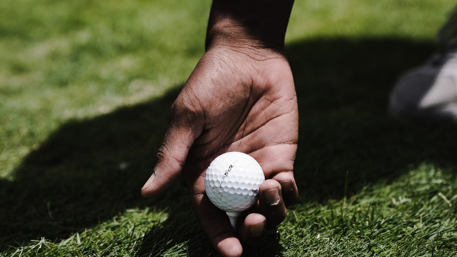 Fotografia em plano fechado que mostra a mão de uma pessoa com a palma voltada para a câmera, próxima a um gramado e com os dedos indicador e médio sob uma bola de golfe no tee; ao fundo, vê-se a sombra do golfista. Foto: Pxhere.