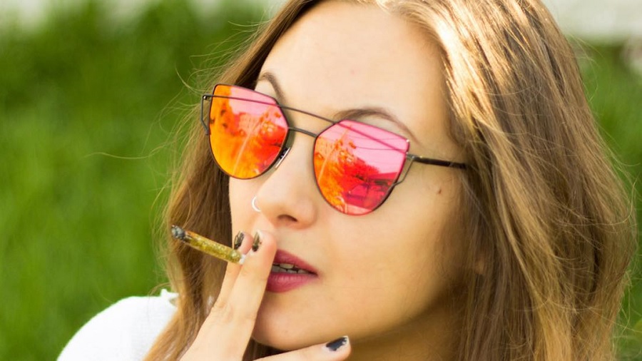 Fotografia que mostra a face de uma mulher que usa óculos com lentes vermelhas espelhadas e segura um baseado (cigarro de cannabis) à boca e um fundo desfocado de vegetação. Foto: Sibeckham | Flickr.