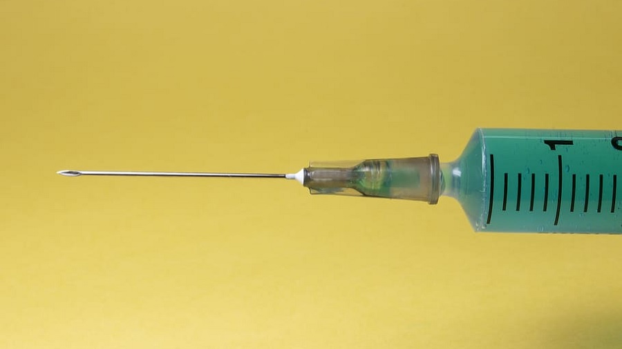 Fotografia que mostra a ponta de uma seringa com agulha, preenchida com uma substância verde, na horizontal e um fundo amarelo. Foto: Pixabay.