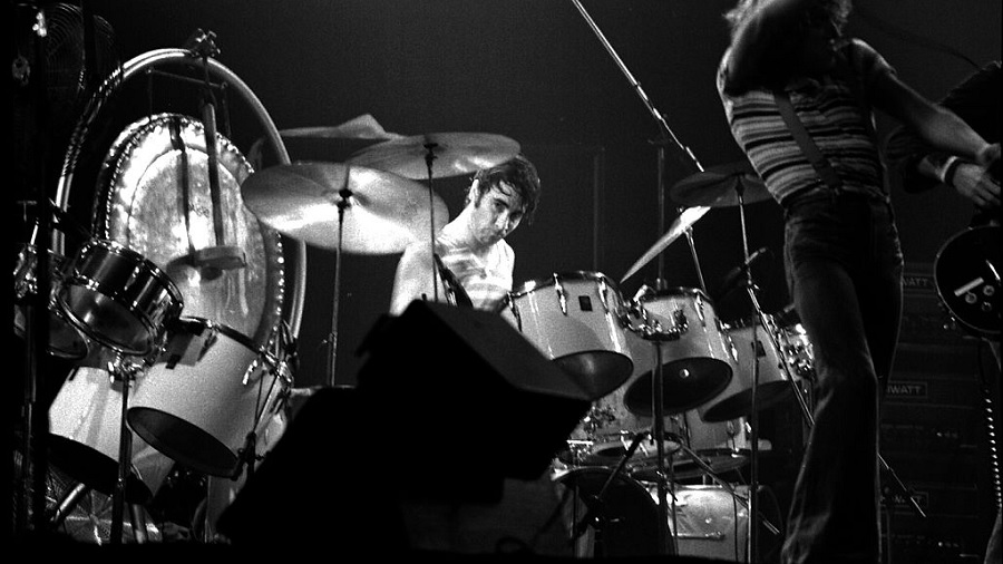 Fotografia em preto e branco do The Who que mostra Roger Daltrey, na parte direita, e, ao centro e em foco, Keith Moon tocando a bateria e olhando para a câmera. Imagem: Jean-Luc | Flickr.