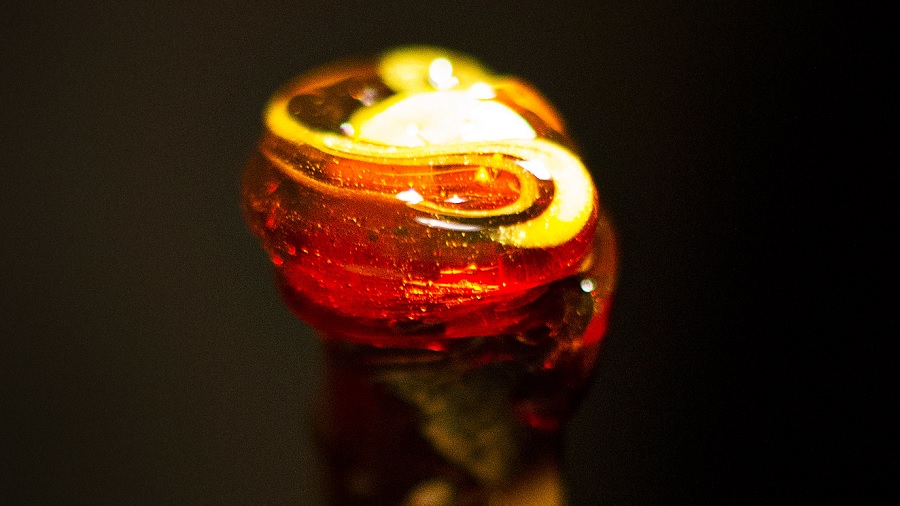 Fotografia em plano fechado que mostra uma porção de concentrado de cannabis tipo BHO, parecido com mel, em torno da ponta de uma haste e sob o foco da luz, e um fundo escuro. Imagem: Andres Rodriguez | Flickr.