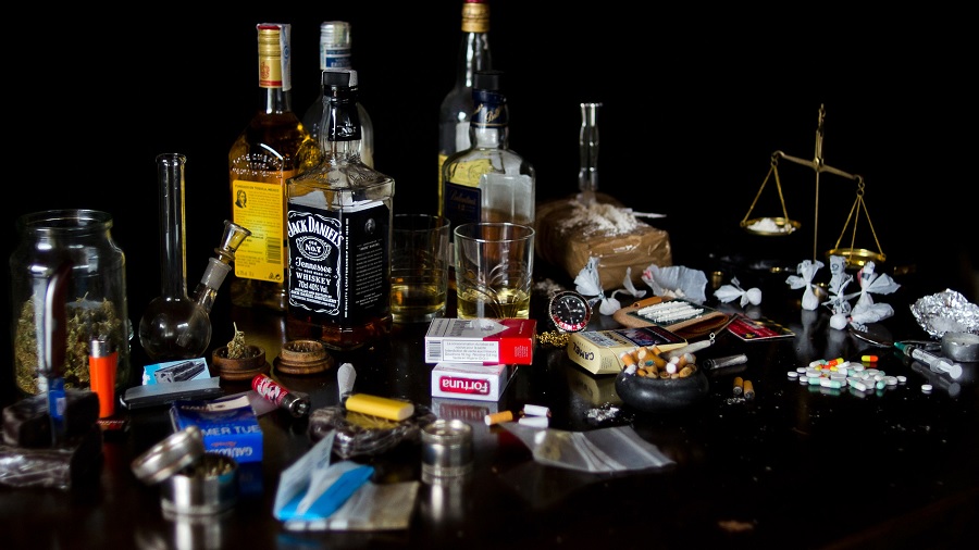 Foto que mostra uma superfície de madeira escura repleta de substâncias (drogas), como garrafas de bebidas, embalagens de cigarros, maconha, cocaína, compridos e seringas, além de acessórios, como isqueiros, cinzeiros e sedas. Foto: Cabrera Photo | Flickr. Unifesp