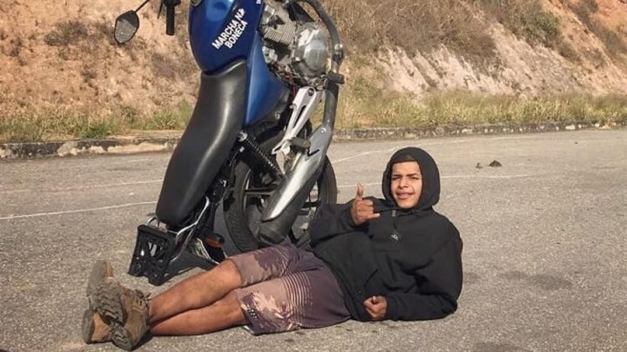 Fotografia que mostra o motoboy Emerson deitado no asfalto e fazendo gesto de ‘hang loose’, ao lado da moto, que está na vertical, apoiada sobre o pneu de trás. Foto: arquivo pessoal.