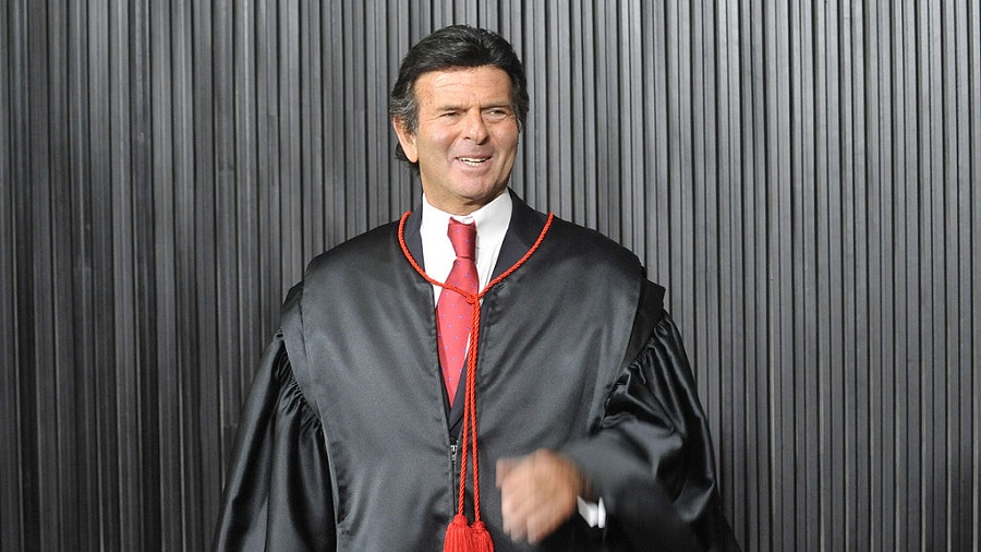 Fotografia que mostra Luiz Fux, da cintura pra cima, usando uma toga com cordonê vermelho e à frente de uma parede estriada de cor cinza-chumbo. Imagem: Jonas Pereira / Agência Senado | Flickr.