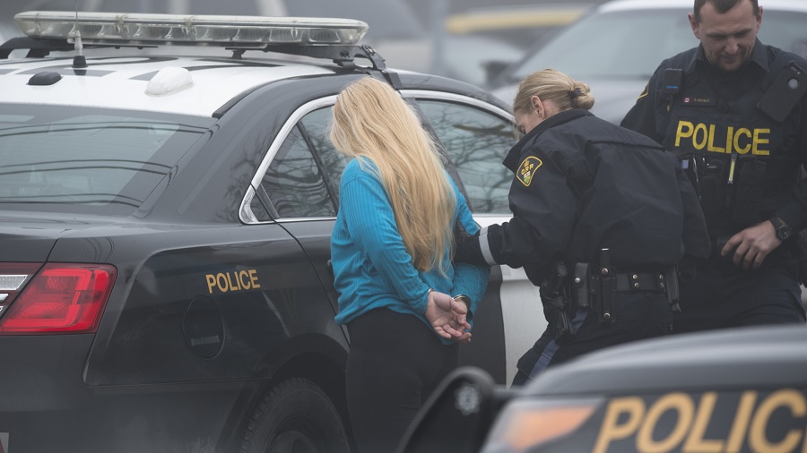 Foto que mostra uma policial levando uma mulher com as mãos algemadas até a viatura, ambas de costas para a câmera, e um policial, na parte direita da imagem. Foto: Polícia Provincial de Ontário.