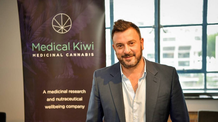 Foto de Aldo Miccio em uma instalação da empresa, onde se vê um pôster institucional da Medical Kiwi com uma foto em preto e branco de folhas da cannabis de fundo, ao lado de uma vidraça, ao fundo. Foto: divulgação.