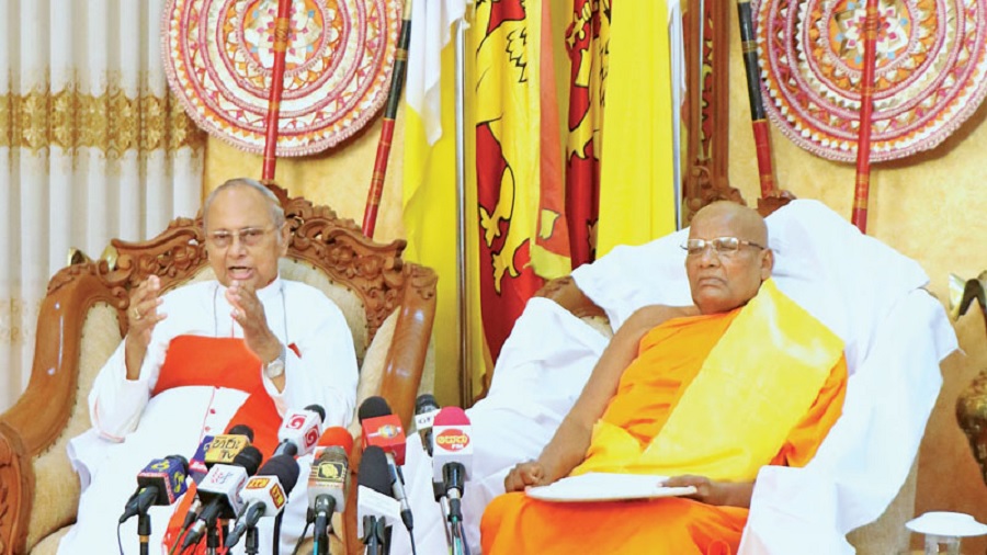Foto de Malcolm Ranjith e Ittapana Dhammalankara Thero, sentados, e diversos microfones, que aparecem na parte inferior da imagem, voltados para eles; ao fundo, se vê uma bandeira do Sri Lanka arriada, entre dois abanadores. Foto: Saman Sri Wedage | Daily News.