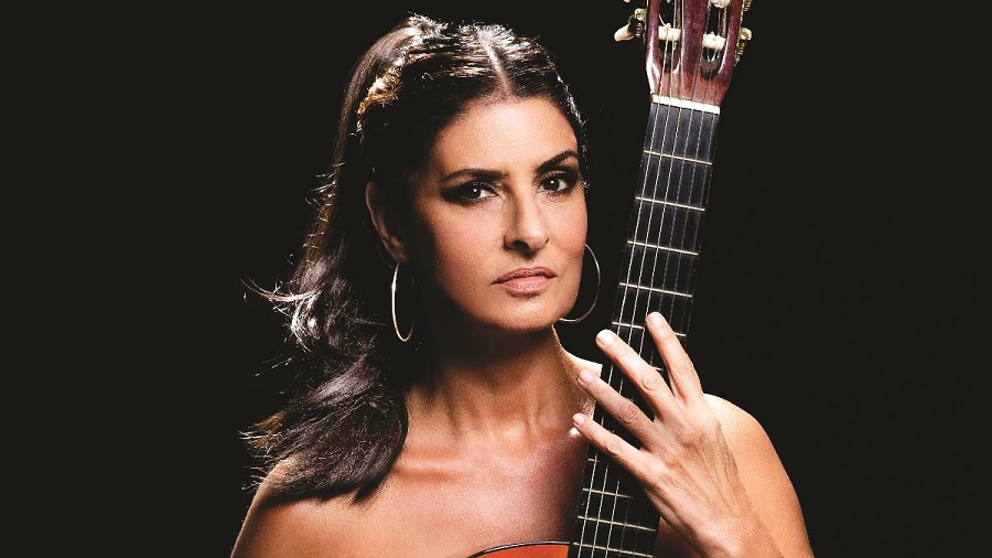 Fotografia em close de Fernanda Abreu segurando um violão em frente ao corpo, do qual aparece o braço, em fundo escuro. Foto: Murilo Alvesso / divulgação.