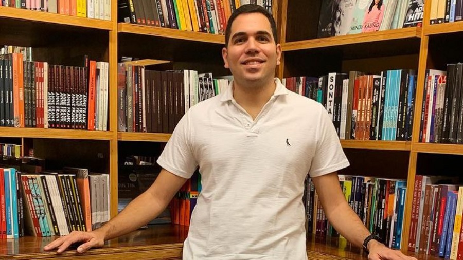 Fotografia que mostra Marcelo Castro encostado em um grande estante de madeira repleta de livros. Foto: reprodução / Instagram.