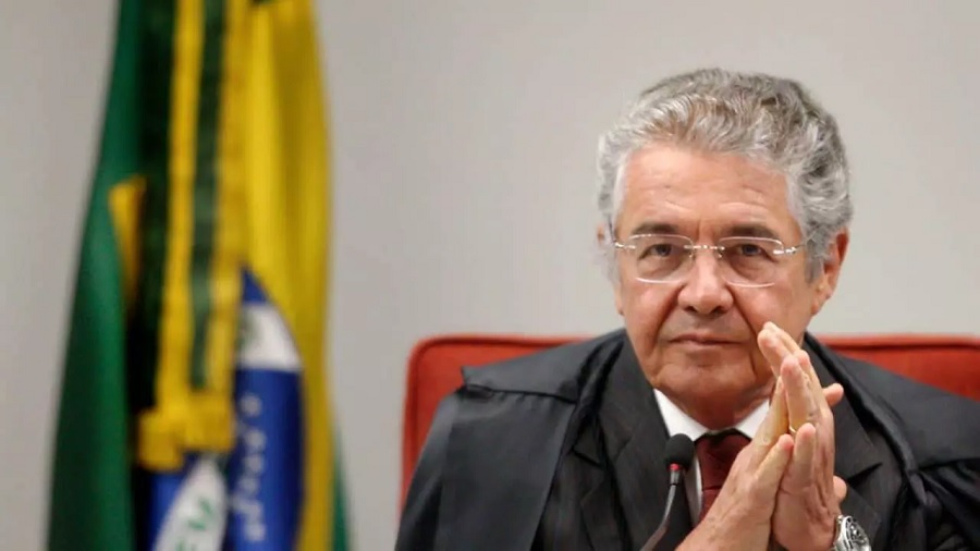 Fotografia que mostra Marco Aurélio Mello, do peito para cima, com as mãos unidas e parte de uma bandeira do Brasil arriada, na parte esquerda da imagem. Foto: Rosinei Coutinho | STF.
