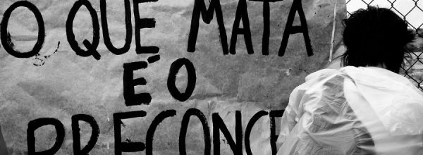Fotografia, em preto e branco, que mostra uma placa com as palavras “O que mata é o preconceito”, fixada em um alambrado, e uma pessoa usando uma capa translúcida, de costas. Foto: Marco Gomes | Flickr.