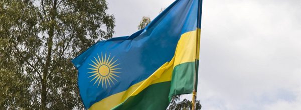 Fotografia que mostra a bandeira de Ruanda flamulando e copas de árvores e um céu com nuvens, ao fundo. Imagem: Hjalmar Gislason | Wikimedia Commons.