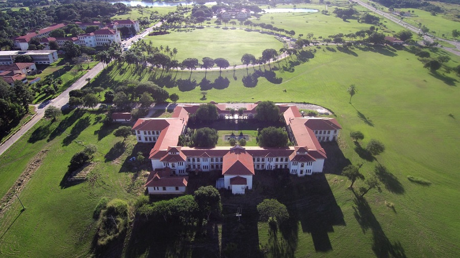 Fotografia aérea do Instituto de Química da UFRRJ, no câmpus de Seropédica, em um momento quando o sol incide do fundo e o prédio e as várias árvores ao redor produzem longas sobras no gramado. Imagem: ilsDrone.