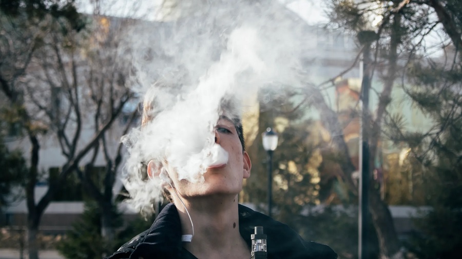 Fotografia que mostra, em primeiro plano, uma pessoa expelindo uma nuvem de vapor e parte do vaporizador, e uma rua arborizada ao fundo. Imagem: Ruslan Alekso | Pexels.