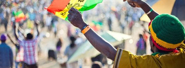 Fotografia que mostra uma pessoa negra de costas e segurando uma pequena bandeira etíope com os braços estendidos e várias pessoas em um ambiente aberto, ao fundo, desfocado. Imagem: Leafly.