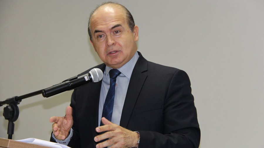 Fotografia de Domingos Sávio de Barros Arruda que, vestido com paletó cinza-escuro e gravata azul, gesticula enquanto fala ao microfone, próximo a uma parede de cor bege.