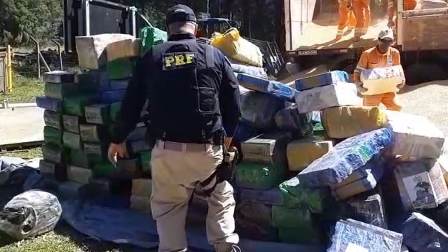 Fotografia de um agente da polícia rodoviária federal de costas, enquanto observa um amontoado de fardos de maconha que são descarregados de um caminhão carregado também com milho. Foto: divulgação / PRF.
