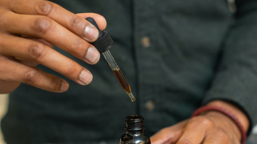 Foto que mostra a mão de uma pessoa segurando um conta-gotas contendo óleo de cannabis de cor marrom, acima de um frasco âmbar, do qual aparece a parte de cima, e parte de seu corpo ao fundo, vestido com uma camisa cinza-escuro. Imagem: R+R Medicinals | Unsplash.