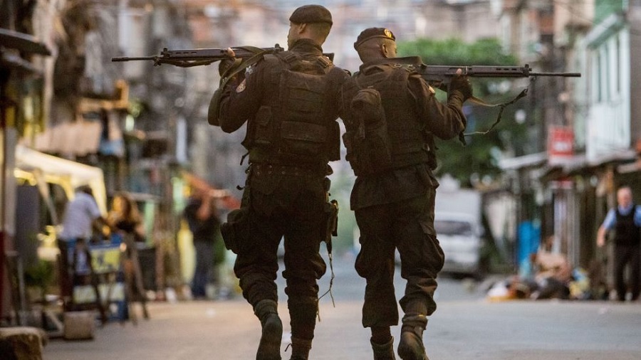 Foto que mostra dois policiais do BOPE, de costas, andando pelas ruas de uma favela do Complexo da Maré, no Rio de Janeiro, enquanto apontam rifles para as laterais da imagem. Crédito: Leonardo Wen.