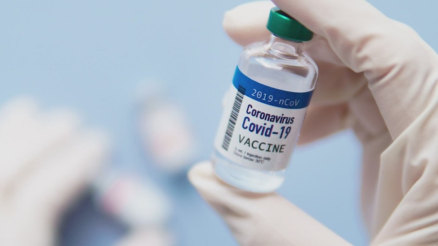 Fotografia de um frasco da vacina contra a Covid-19, com rótulo azul e branco e tampa verde, sendo segurado por uma mão com luva cirúrgica branca, e uma superfície azul-claro onde estão outros dois frascos e a outra mão da pessoa, ao fundo, desfocado. Imagem: KHOU 11.
