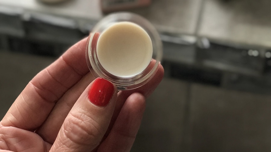 Foto de um pequeno recipiente redondo transparente contendo creme de cor pérola sendo segurado com a parte de cima voltada para a câmera (detalhe para o esmalte vermelho no polegar). Imagem: Becky Fantham | Unsplash.