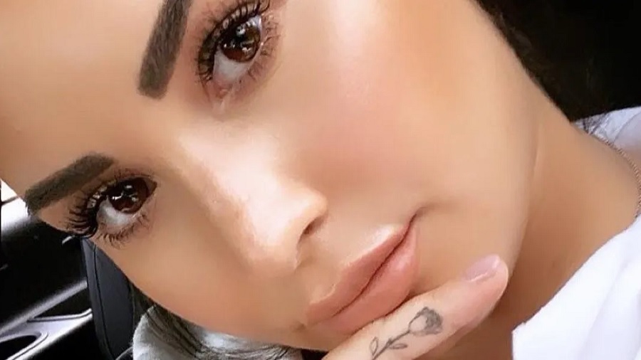 Fotografia em close de Demi Lovato, que aparece com a cabeça inclinada para o lado e o dedo, tatuado com uma flor, próximo à boca. Imagem: reprodução / Instagram.