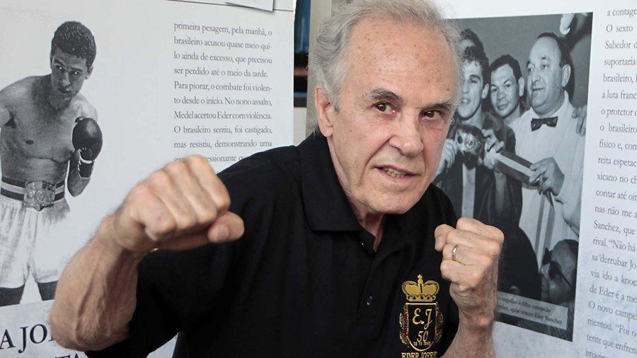 Fotografia de Eder Jofre usando uma camisa preta com seu brasão de 50 anos de cor dourada, em posição de combate, à frente de pôsteres com fotos antigas do boxe. Imagem: divulgação.