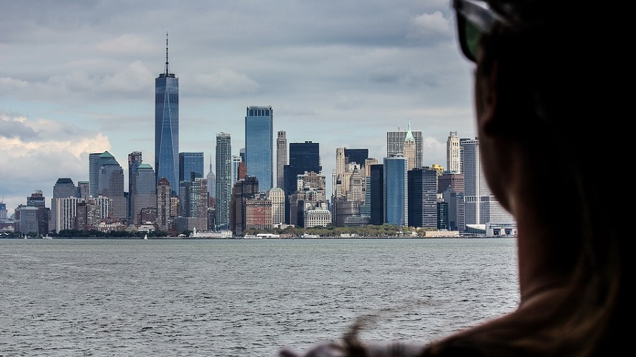 Foto que mostra parte do ombro e cabeça de uma pessoa, que aparece de costas no lado direito do quadro, no primeiro plano, e a Ilha de Manhattan, ao fundo, após as águas do Atlântico. Imagem: Zsubio | Pexels.