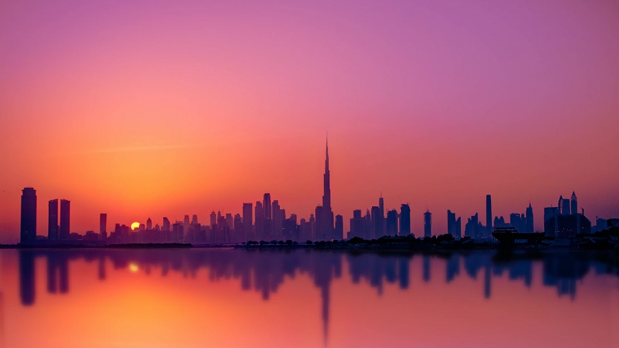 Fotografia que mostra uma vista geral da cidade de Dubai, onde a silhueta dos prédios, com o arranha-céu Burj Khalifa ao centro, e um céu em tons de rosa e laranja são refletidos nas águas do Golfo Pérsico. Foto: Mohammed Nasim | Unsplash.