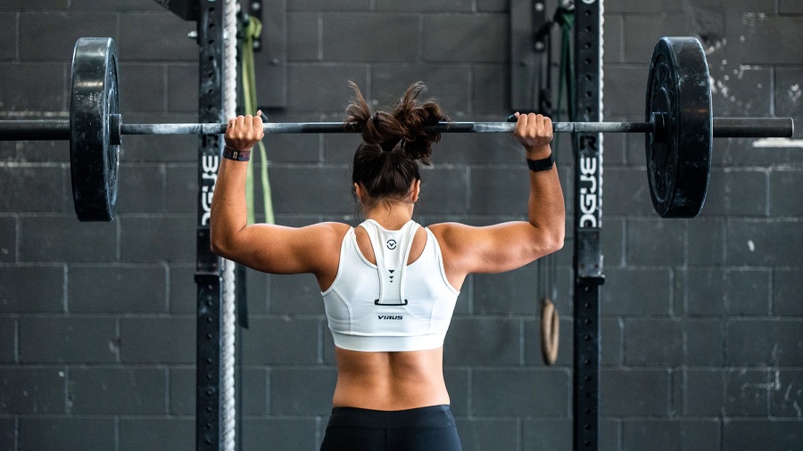 Fotografia que mostra uma mulher de costas segurando uma barra com duas grandes anilhas, uma em cada ponta, acima da cabeça, de frente ao suporte de barras que está fixado em uma parede de blocos cinza. Foto: John Arano / Unsplash.