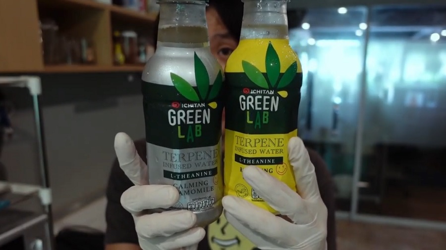 Fotografia que mostra uma pessoa segurando duas garrafas da bebida da Ichitan, uma com rótulo em fundo cinza e outra amarelo, onde se vê a marca “Green Lab” sobre uma tarja verde e o logo de folha de cannabis que traz o nome da empresa no lugar de um dos folíolos, na Tailândia. Imagem: Youtube / Lower100THB.