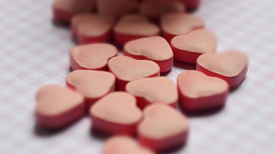 Fotografia de uma porção de comprimidos rosas em formato de coração sobre uma superfície xadrez branca e rosa-claro. Imagem: Pexels / Dear W.