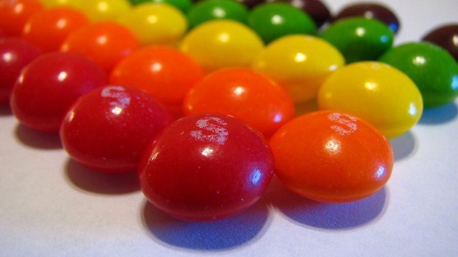 Fotografia que mostra uma porção de balas Skittles dispostas em fileiras por cor, vermelho, laranja, amarelo e roxo, na diagonal, em uma superfície branca. Foto: Flickr | Amber.