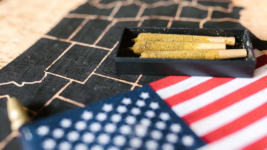Fotografia mostra uma caixinha preta retangular com três baseados (cigarros de maconha) empanados em seu interior, sobre uma superfície de madeira que tem um mapa dos EUA de cor preta, e a parte de cima da bandeira do país, no canto inferior direito da imagem. Foto de RODNAE Productions no Pexels. veteranos
