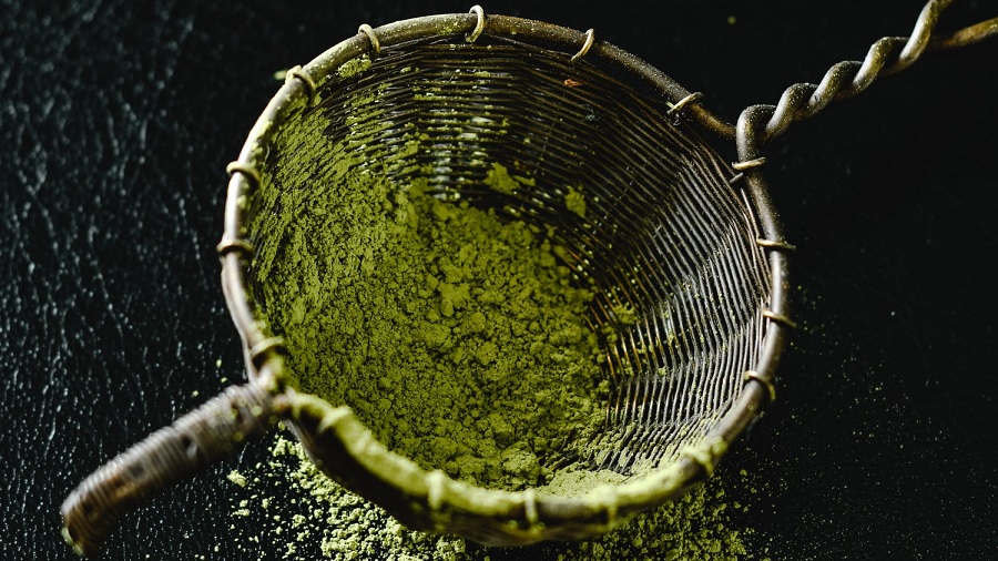Fotografia de uma escumadeira metálica contendo um pouco de farinha verde, que passa pelas frestas e cai sobre uma superfície preta texturizada. Imagem: Eva Elijas / Pexels.
