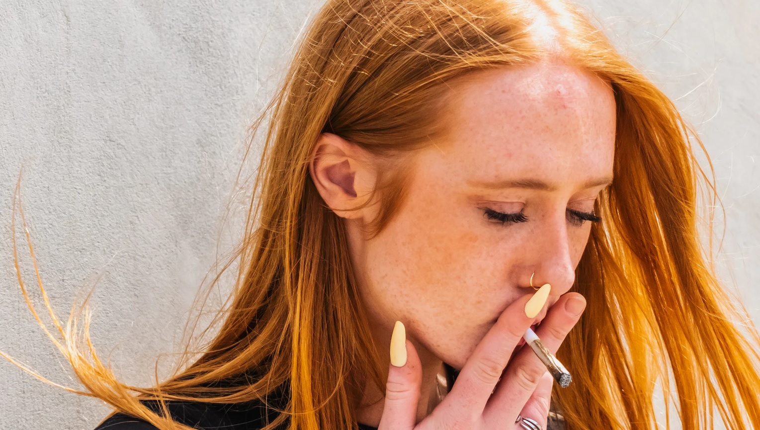 Foto, em close e meio perfil, de uma mulher de cabelo ruivo que segura um cigarro de maconha aceso à boca, entre os dedos indicador e médio, mostrando as unhas compridas amarelas no polegar e indicador. Fotografia: Alex Woods | Unsplash.
