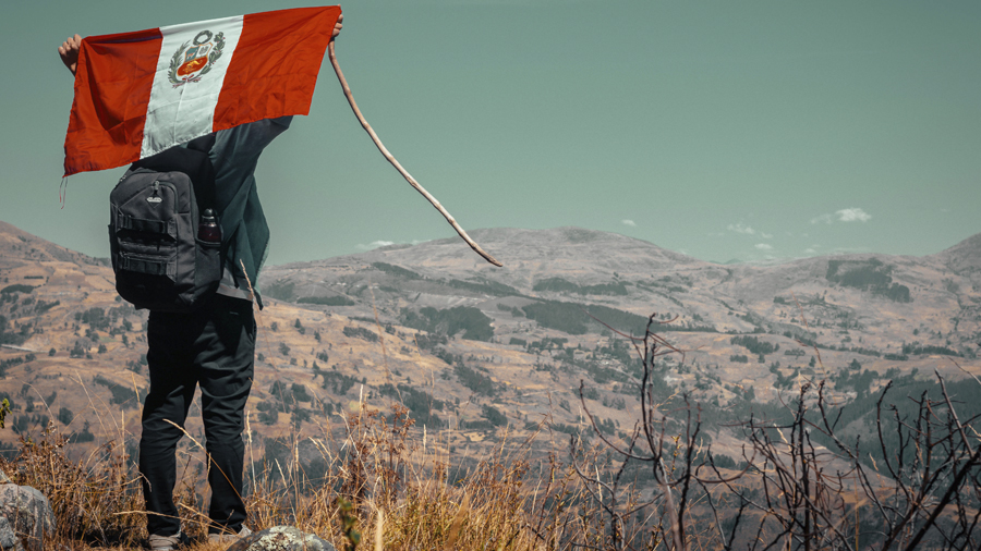 Fotografia mostra uma pessoa carregando mochila e cajado com a bandeira do Peru esticada sobre os ombros, em um mirante de frente para o horizonte onde se vê as montanhas de Apurimac. Crédito: Carlos Ruiz Huaman | Unsplash.
