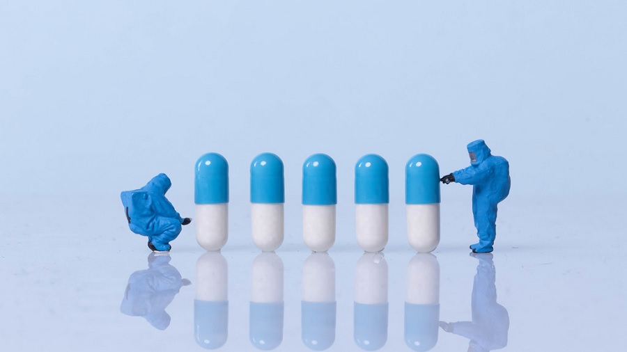 Fotografia mostra uma fila de cápsulas de cor azul e branca, em pé, no meio de duas miniaturas de pessoas com uniforme azul, em uma superfície azul-claro que se mistura ao fundo da imagem. Crédito: Marco Verch | Flickr.